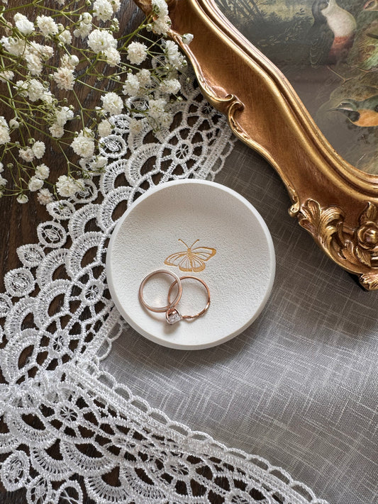 white clay ring dish with gold butterfly design in center in flat lay photo with babies breath and gold frame